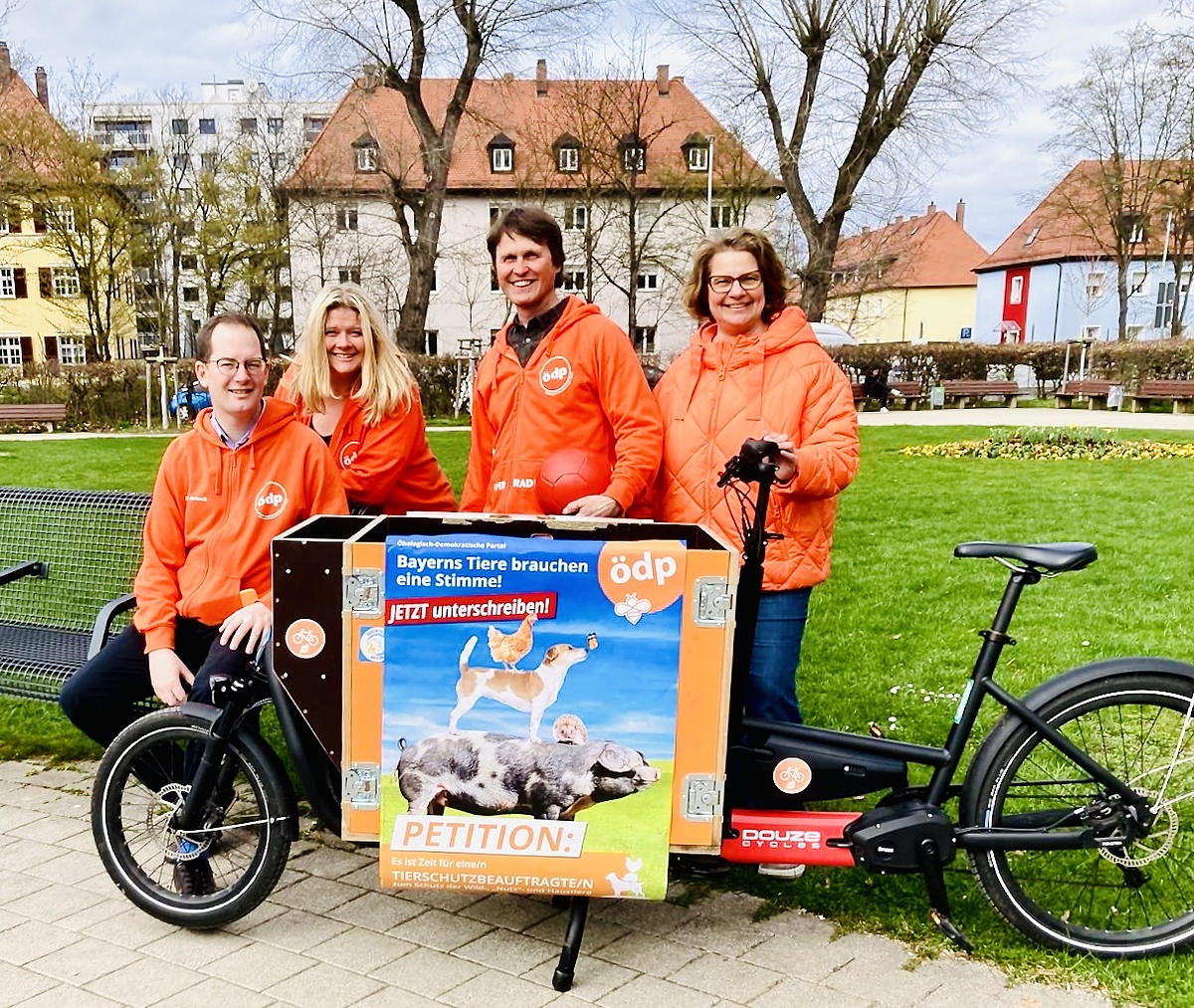 Direktkandidaten 2023 Das Foto zeigt vier erwachsene Personen in oranger Kleidung (mit ÖDP-Logo) und ein Lastenrad, im Hintergrund eine Wiese mit Blumen (Ohmplatz) sowie Bäume und Häuser. Auf dem Lastenrad klebt ein Plakat für die Tierschutz-Petition der ÖDP.